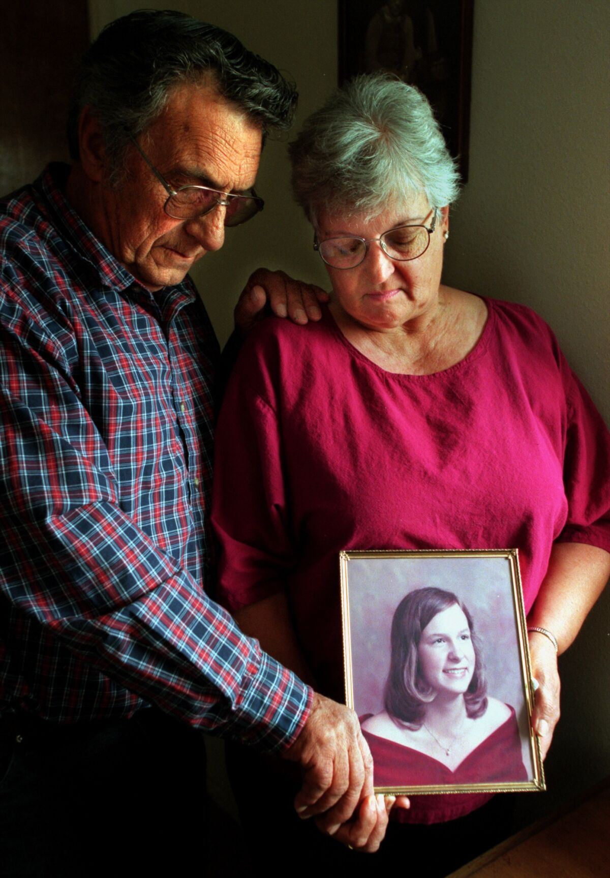 A man and a woman stand close together and both hold a framed picture of a young woman, looking at it.
