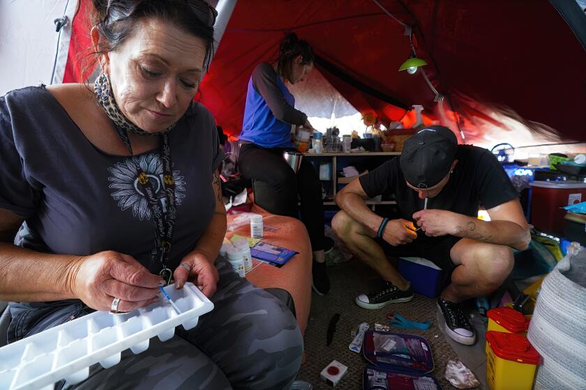 As Billy smokes fentanyl inside his tent at a homeless encampment, Tara Stamos-Buesig (l) test small samples of the drugs