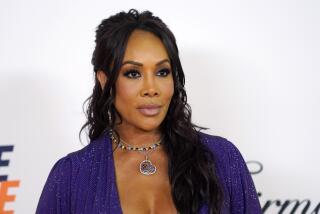 Vivica A. Fox poses in a sparkly purple outfit and chunky heart necklace against a white background.