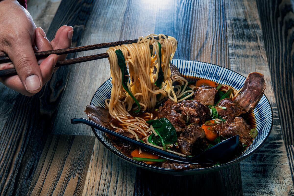 A hand uses chopsticks for a bowl of beef noodle soup