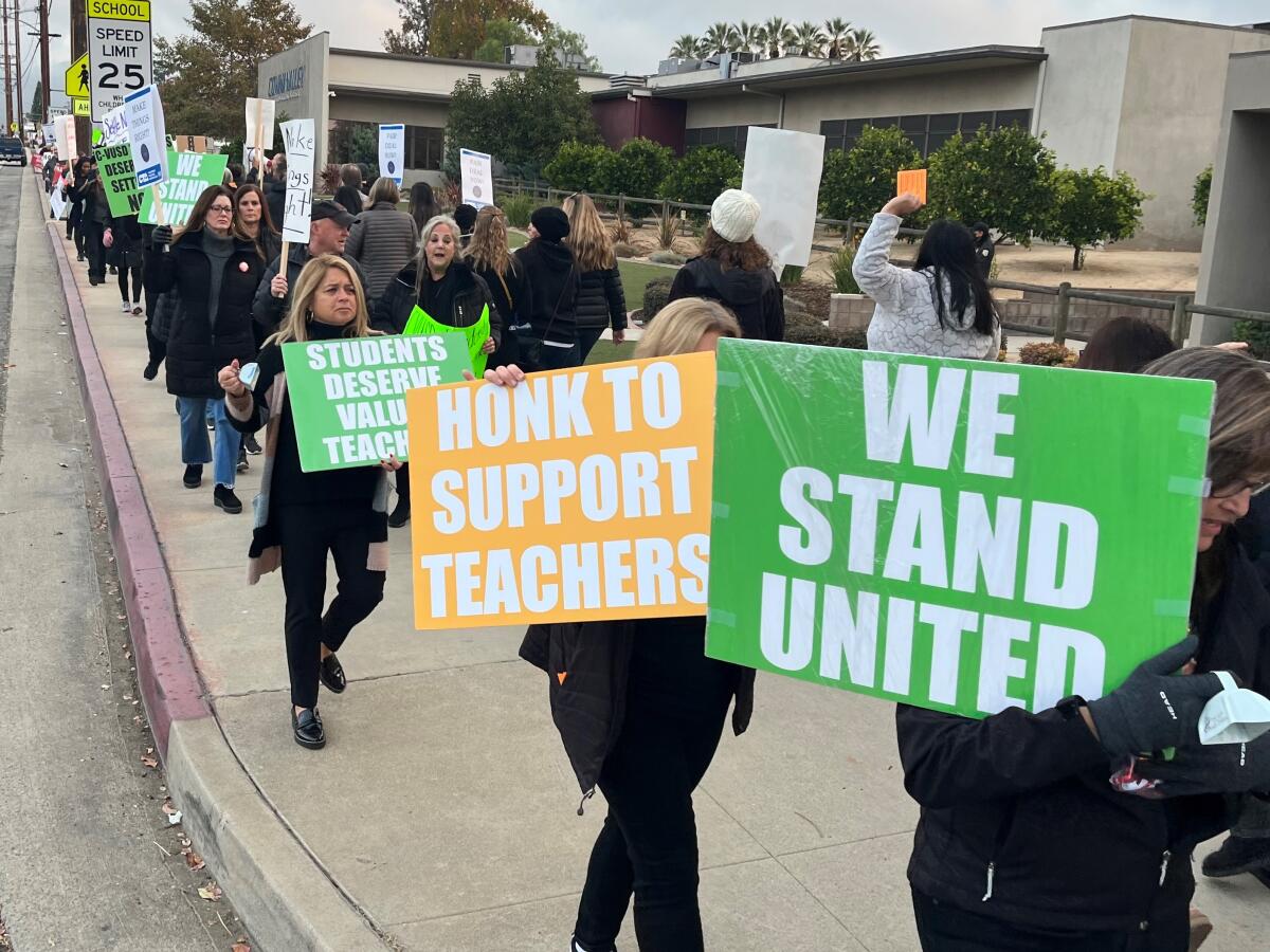 Teachers walk on a sidewalk with signs reading "Honk to Support Teachers" and "We Stand United"