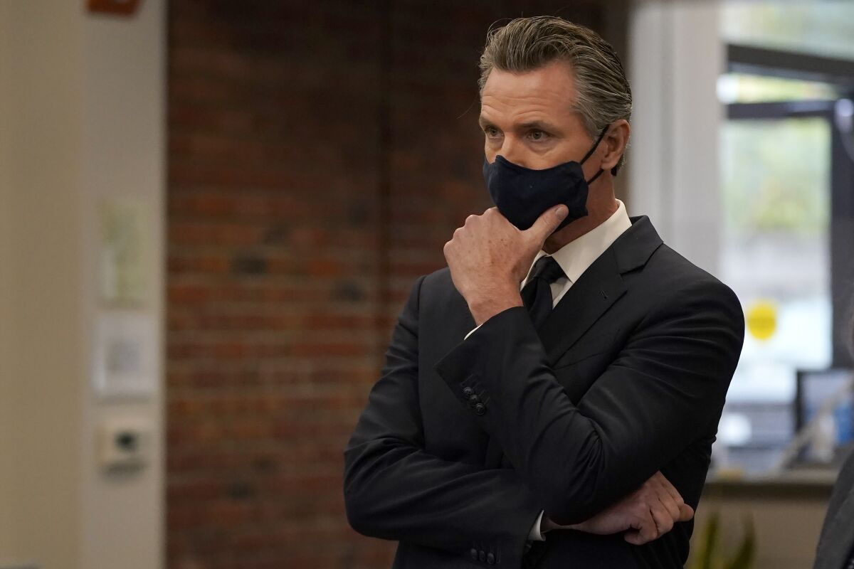 Gavin Newsom, in a mask, stands with his hand on his chin.