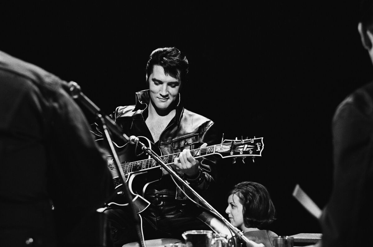 A black and white photo of Elvis Presley looking down at his guitar.