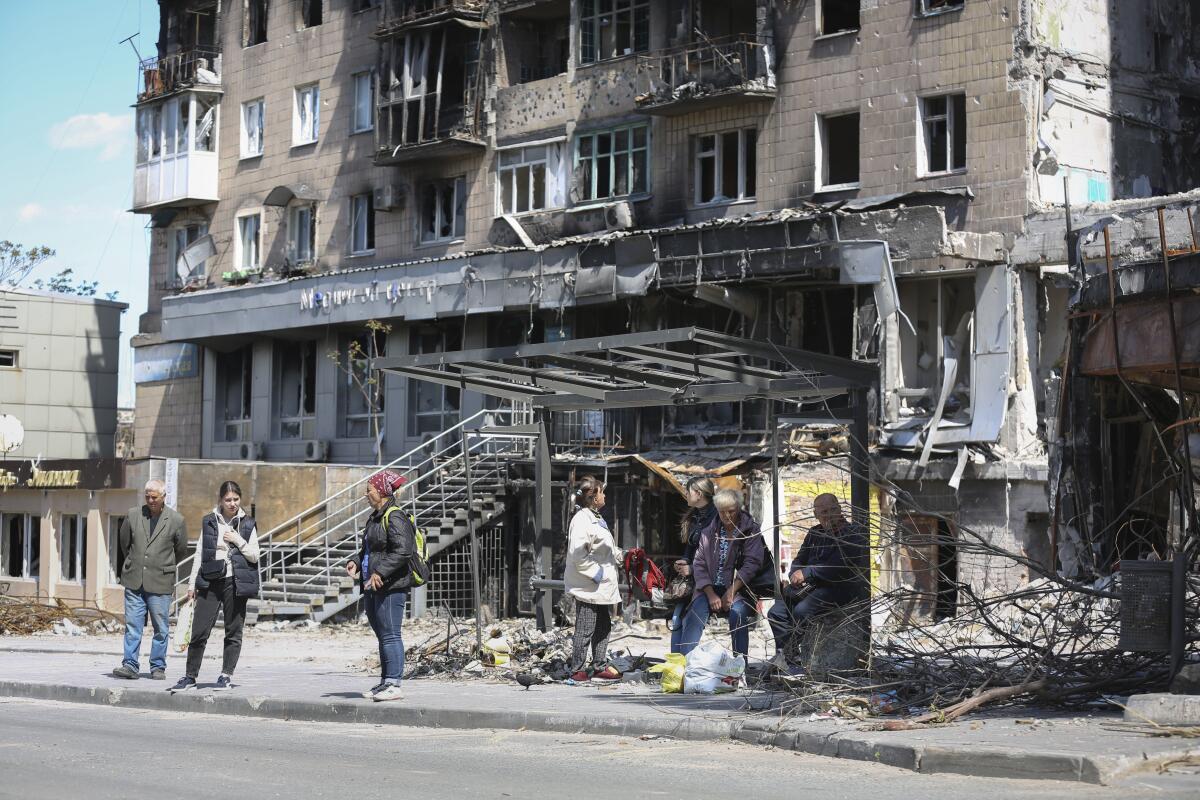 People standing at a bus stop by a damaged building.