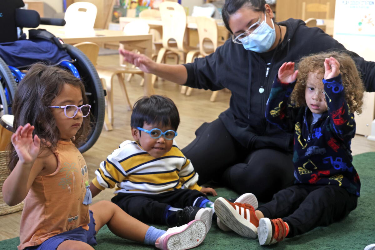 Three preschoolers, two of whom wear glasses, sit on the rug next to their teacher lifting their hands into the air.