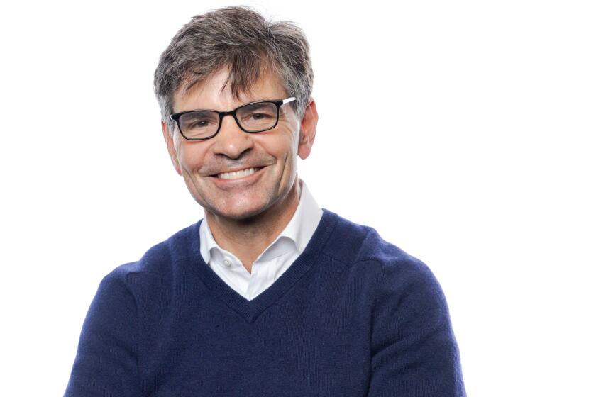 George Stephanopoulos wearing a navy blue sweater and smiling against a white backdrop