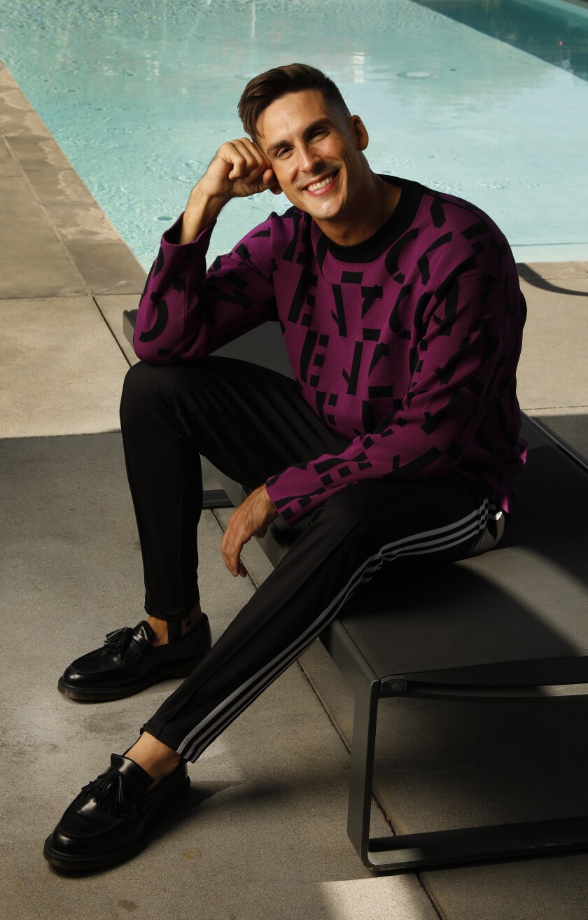 A man in a purple sweater, track pants and black shoes poses by a pool.