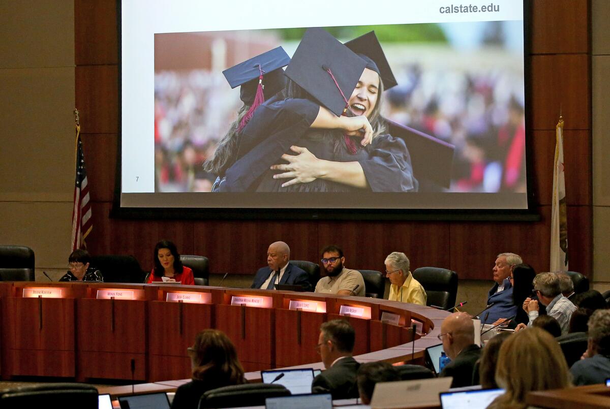 Several people seated around a curved table below a large screen showing students in graduation caps and gowns hugging