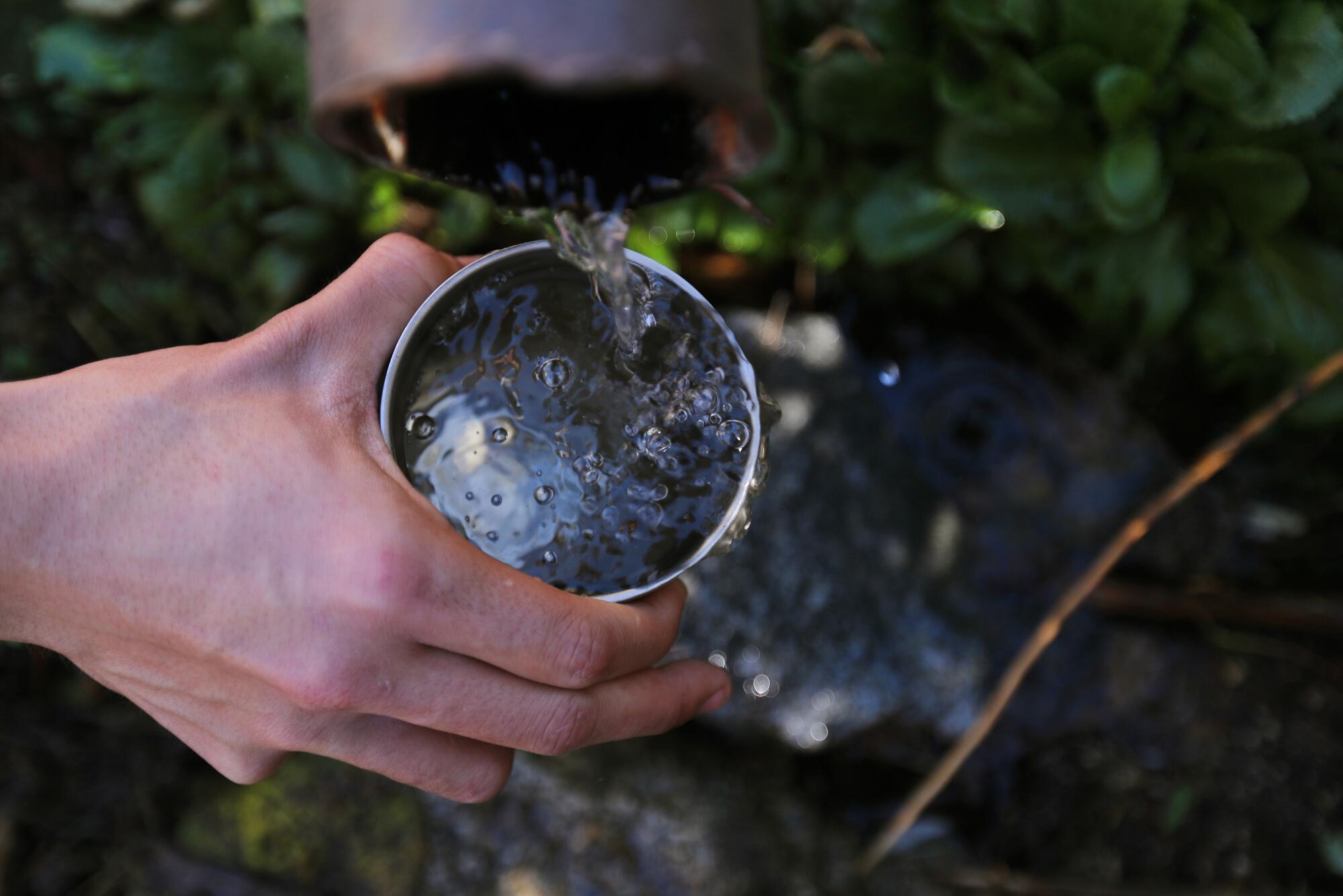 Water flows from a pipe into a metal cup held a hand.