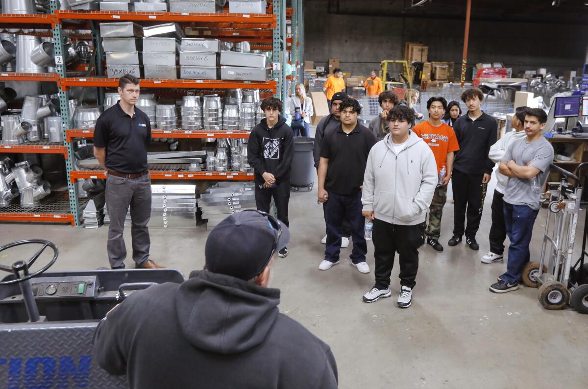 A group of teens stand in a warehouse as a man speaks to them.