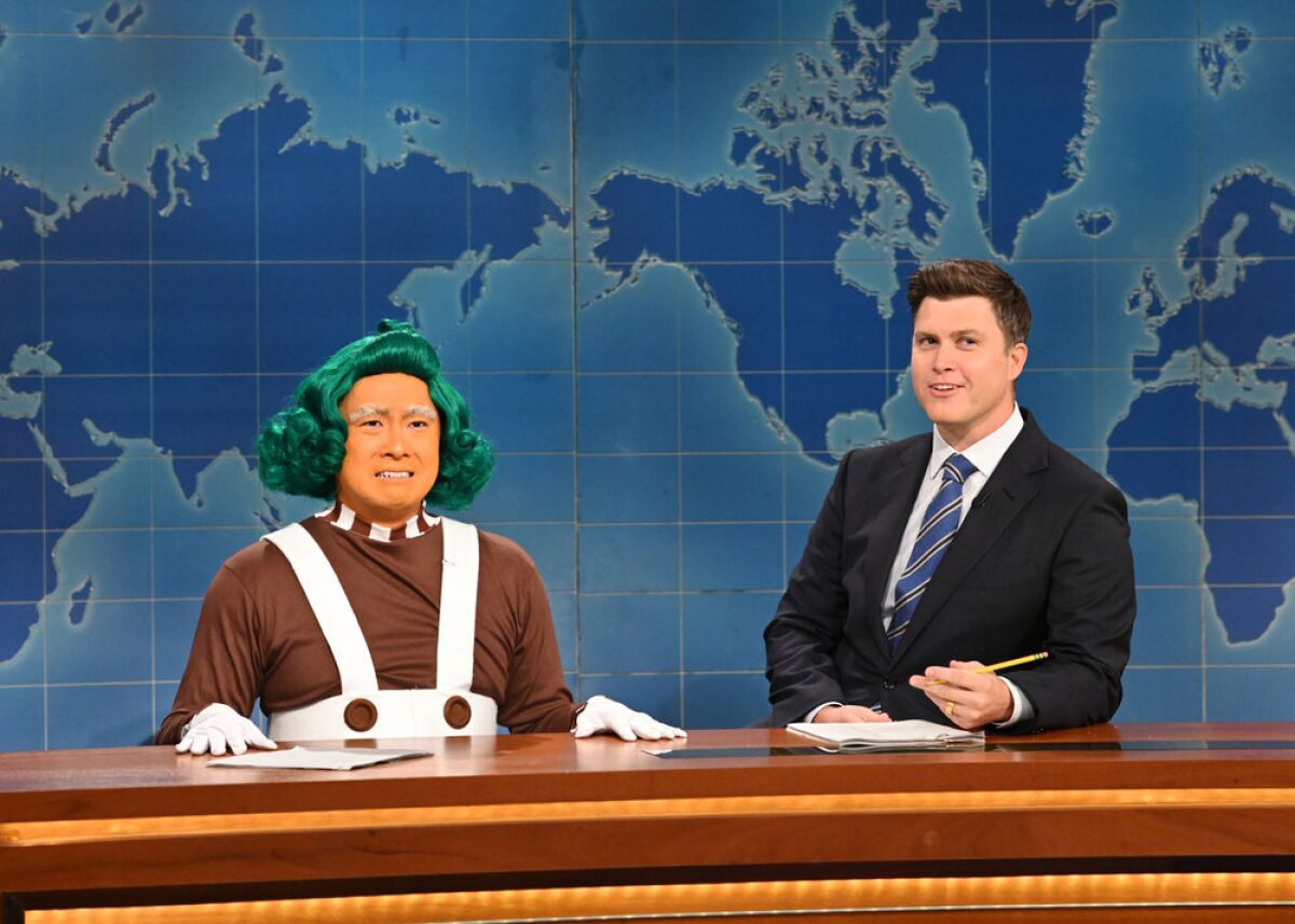 A man wearing orange face makeup and a green wig, sitting next to a man in a suit behind a desk