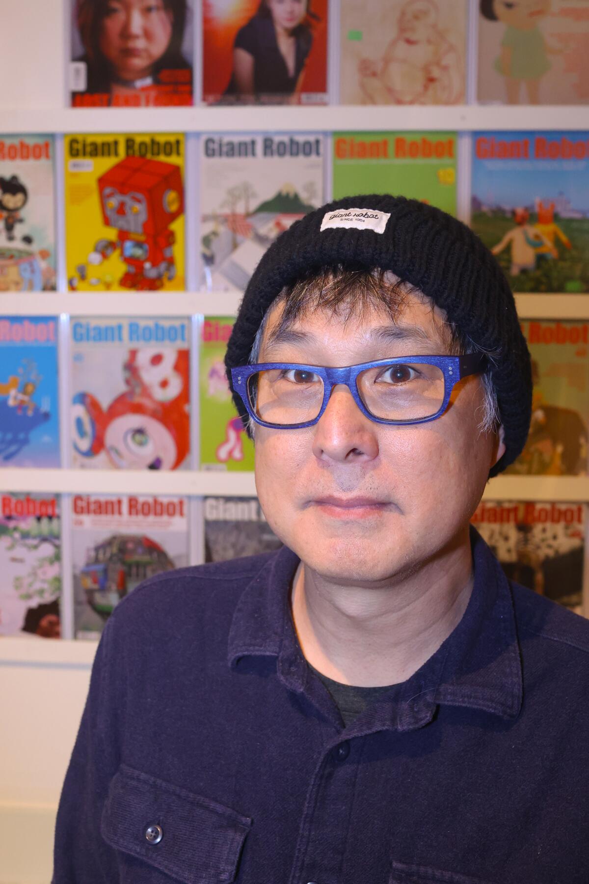 A man wearing glasses and a wool hat stands in front of a display of magazine covers.
