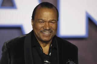 Billy Dee Williams in a dark jacket smiling against a blurry backdrop with large letters
