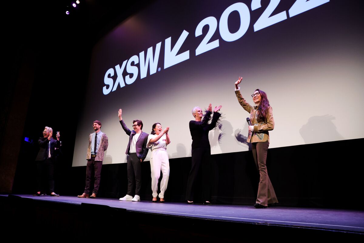 Directors and cast members of 'Everything Everywhere All at Once' stand on a stage in front of a movie screen
