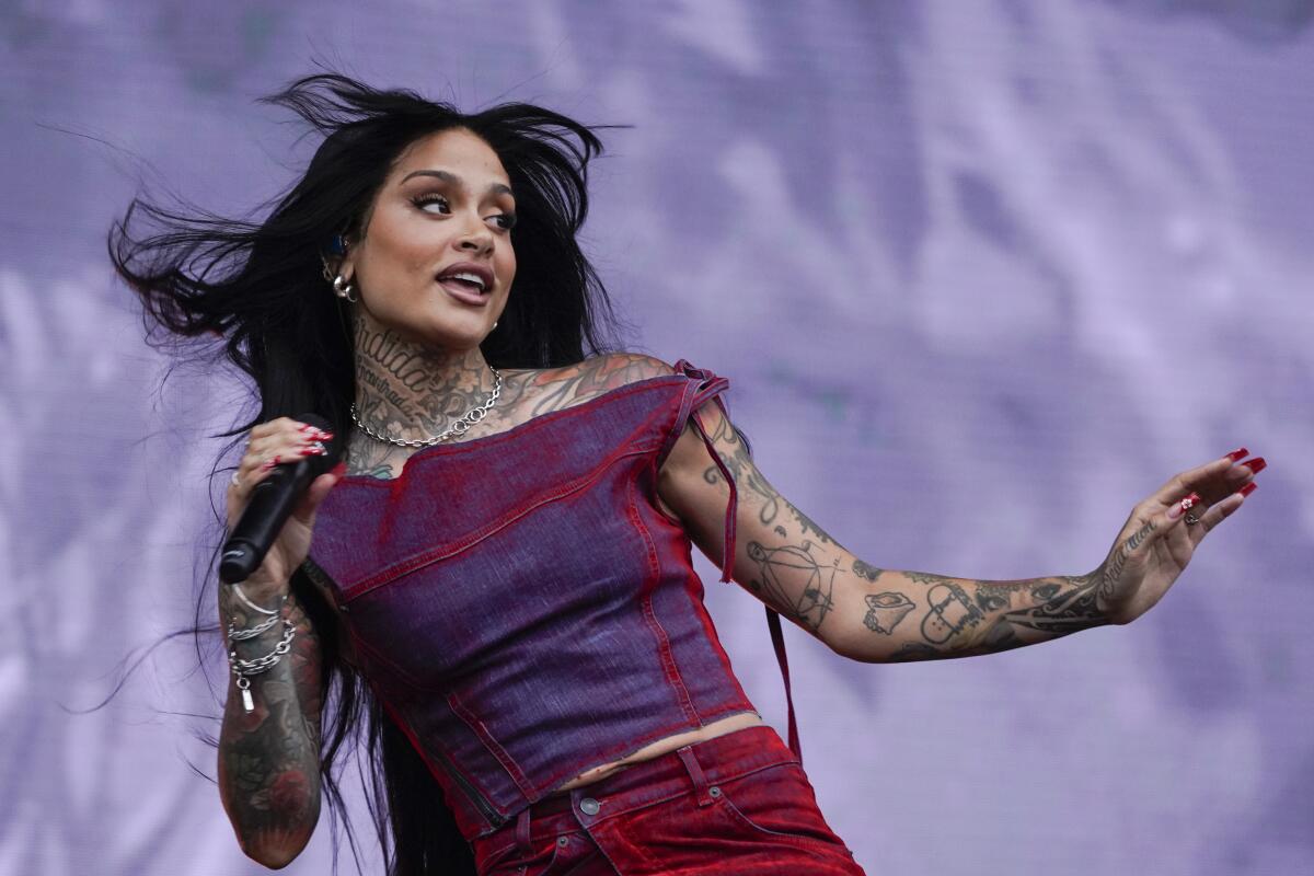 Kehlani on stage holding microphone with other arm held out and her hair blowing in the wind in a purple and red matching set