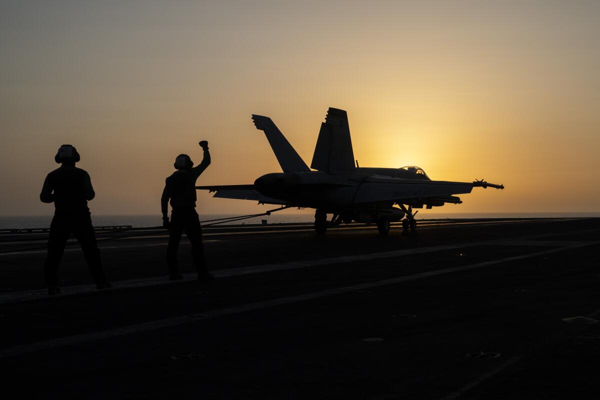 A fighter jet and two personnel are silhouetted on an aircraft carrier. 