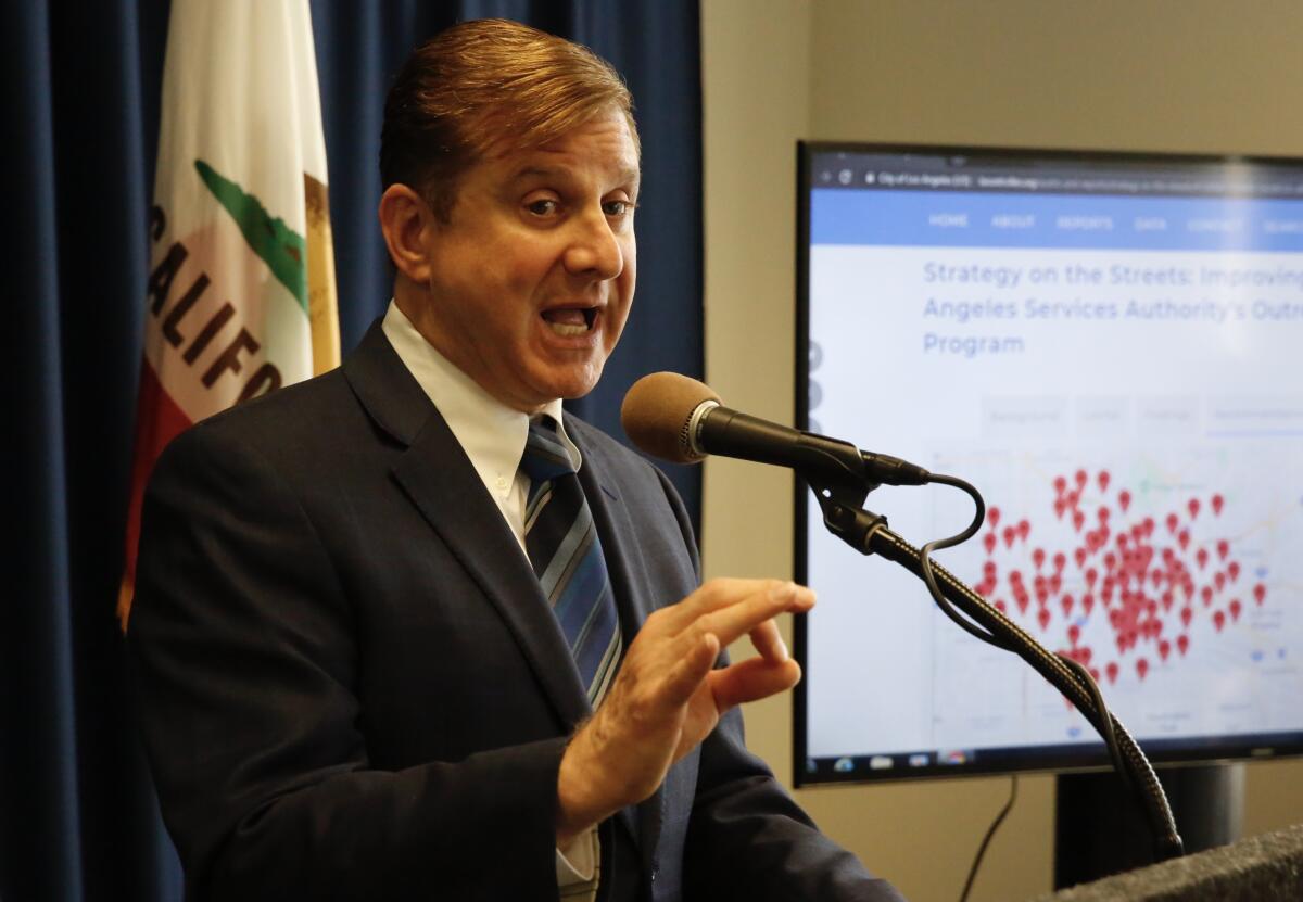 Los Angeles City Controller Ron Galperin gesturing as he speaks into a microphone next to a screen displaying a graphic 