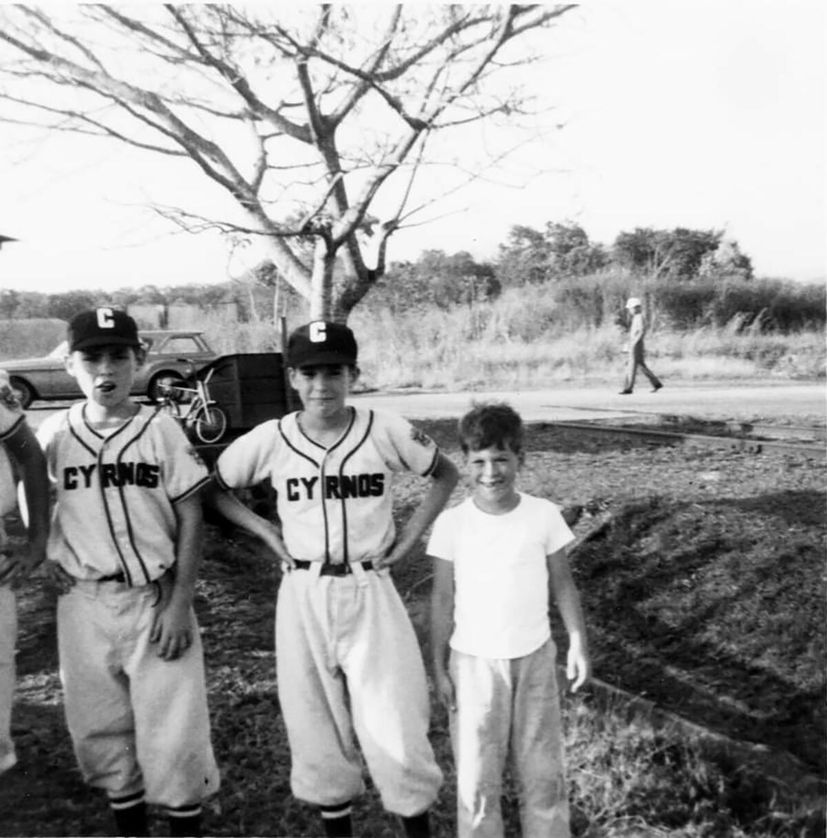 Three boys, two in baseball uniforms, smile in a black-and-white photo.