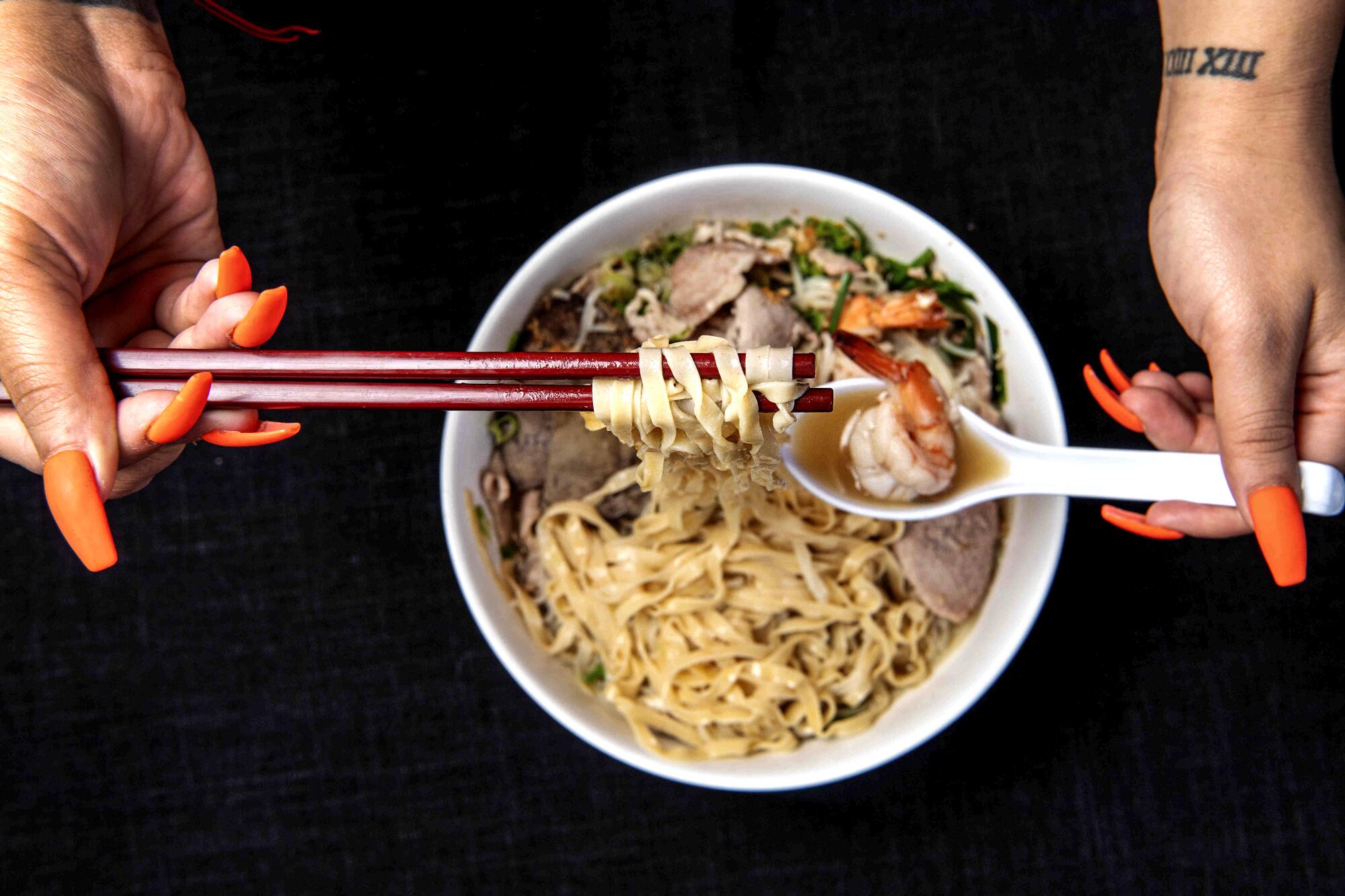 Hands hold chopsticks with noodles and a spoonful of shrimp above a bowl of House Special Noodles.