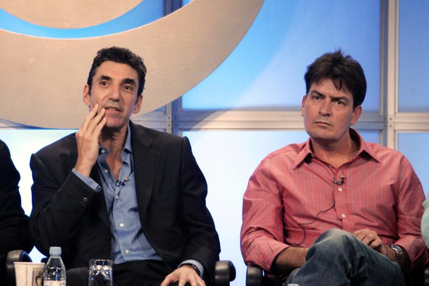 Chuck Lorre in black suit with blue shirt and Charlie Sheen in salmon shirt, seated, talking from stage