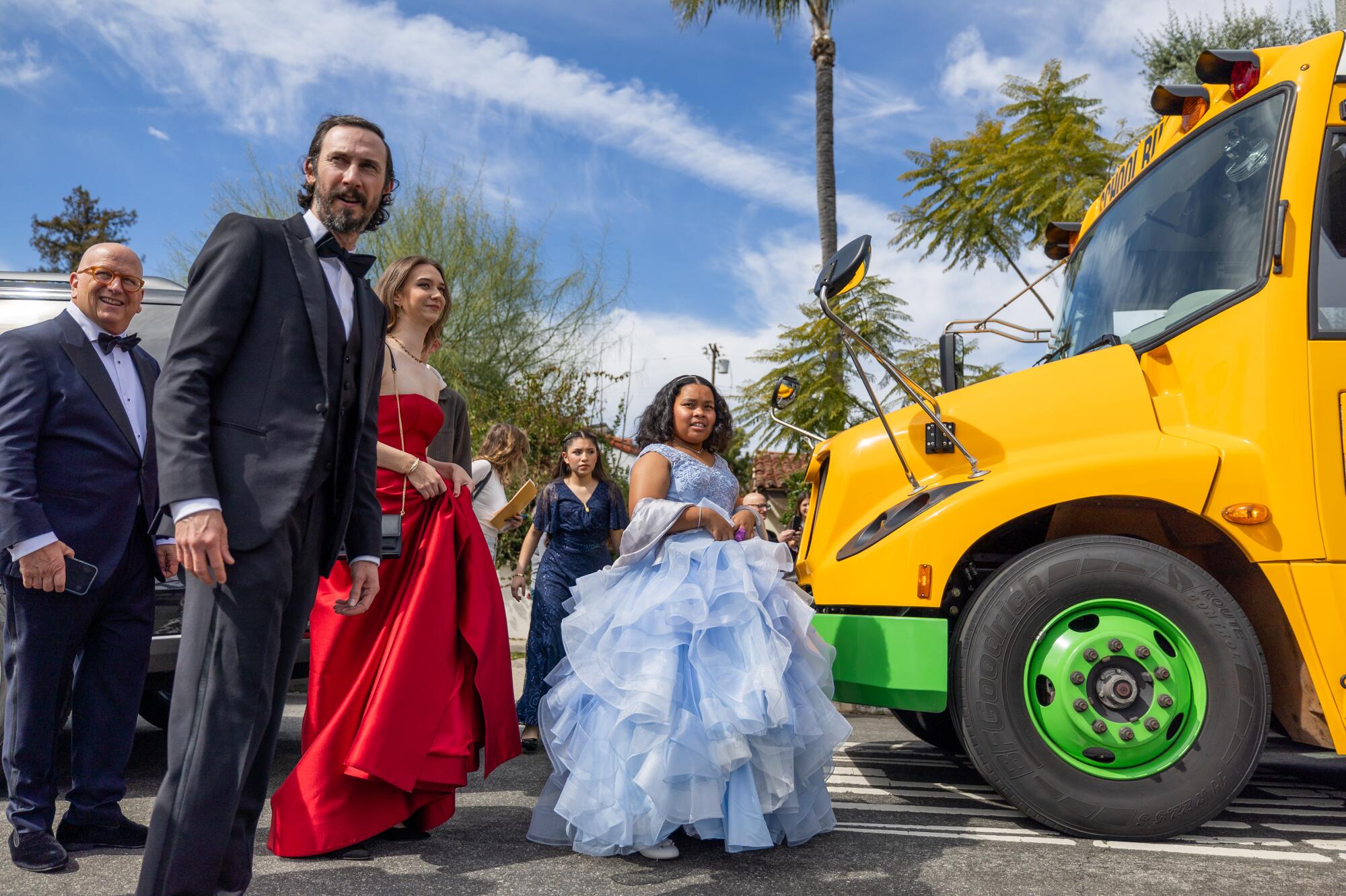 A young girl wearing a frilly light blue dress and surrounded by adults in formalwear boards a school bus.