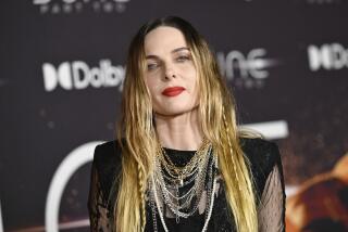 A woman with long blond hair wearing a dark dress and multiple necklaces posing against a dark background