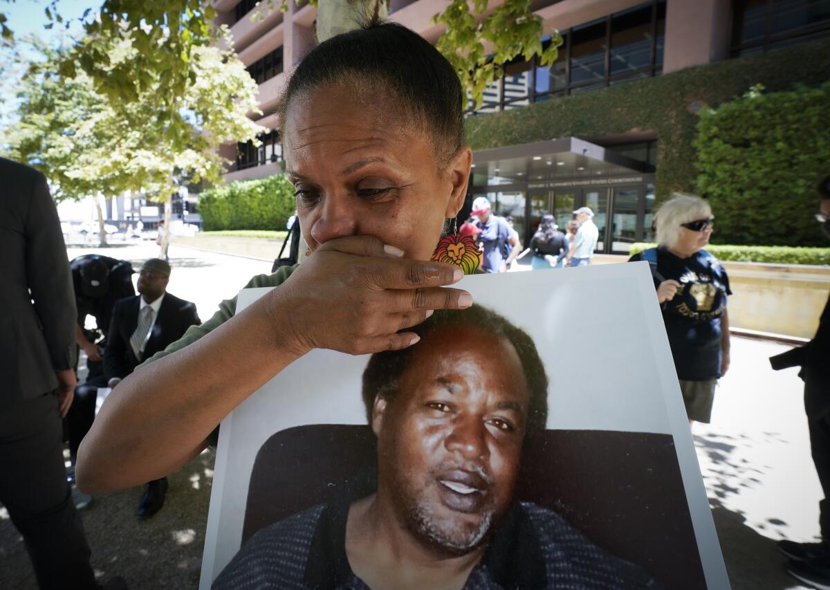 A woman covers her mouth and looks toward the ground as she holds a poster-sized photograph of her deceased brother