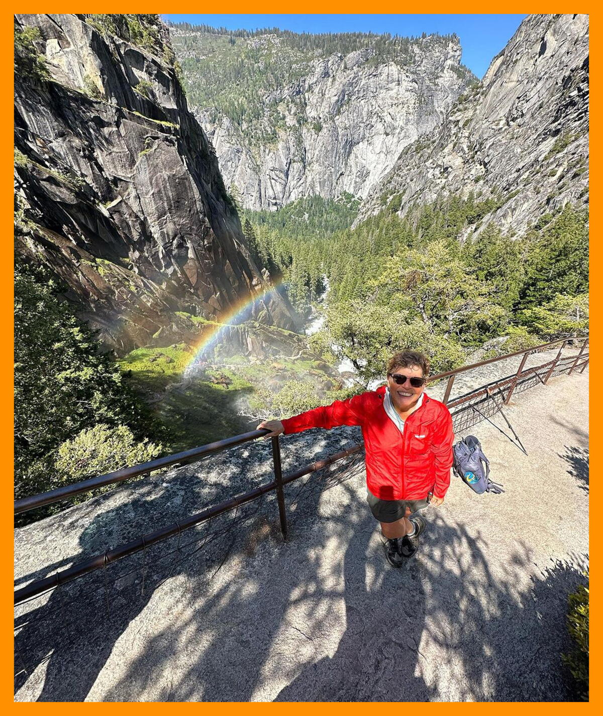 A hiker stands in front of a roaring waterfall with a rainbow forming in the mist