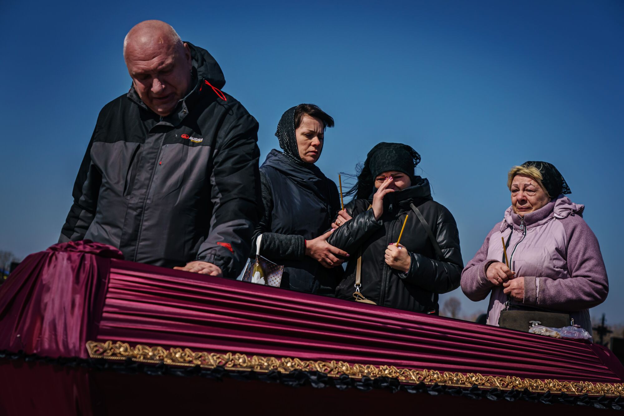 A man places his hand on a cloth-draped casket against a clear blue sky as three women look on.