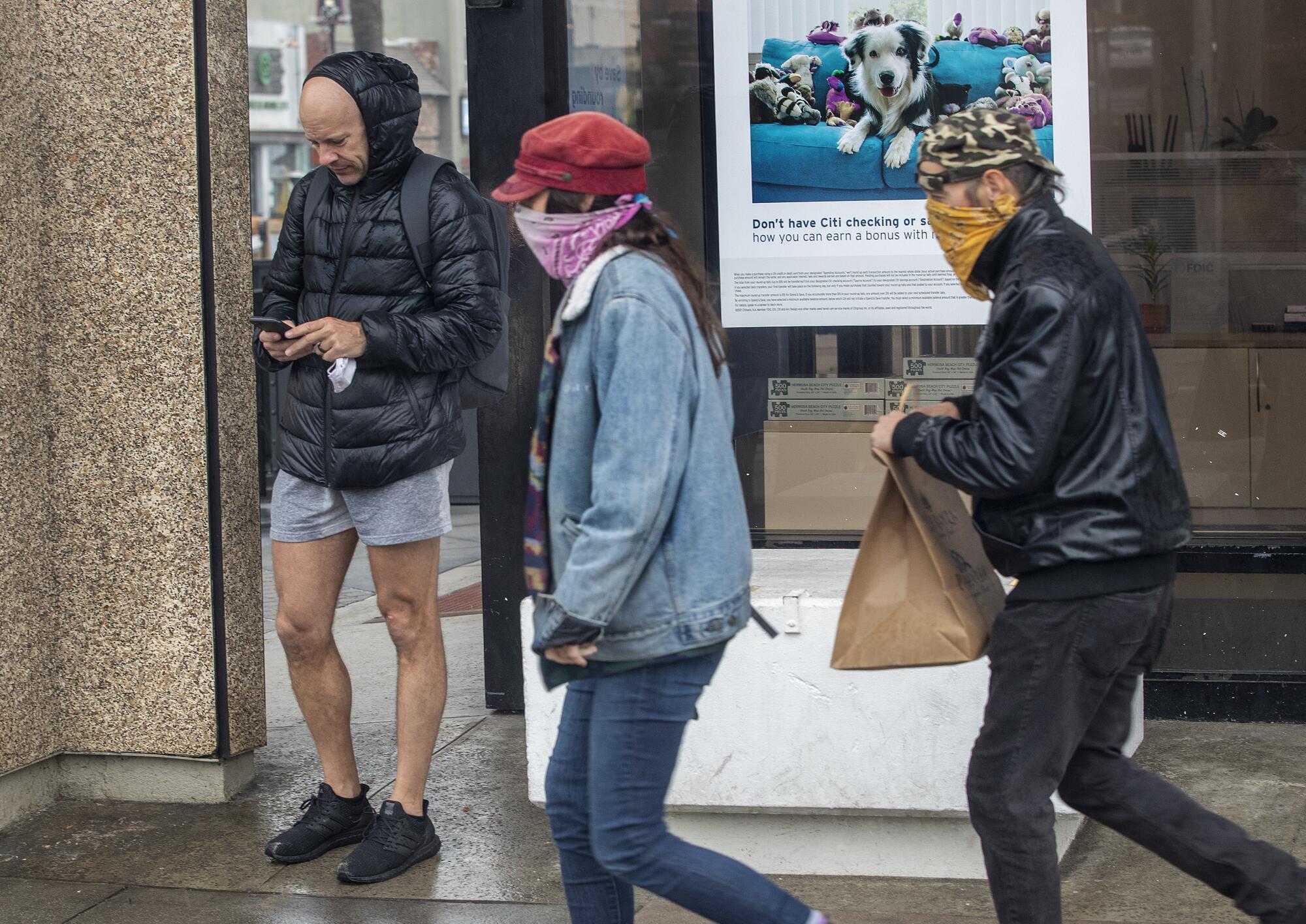 People wearing bandannas as masks walk on a sidewalk next to a man in a coat and short pants