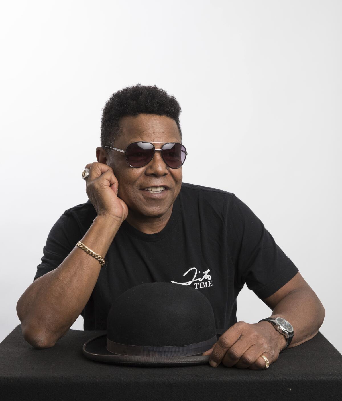 Tito Jackson in a black T-shirt and aviator sunglasses poses with his elbow on a table in front of a black bowler hat