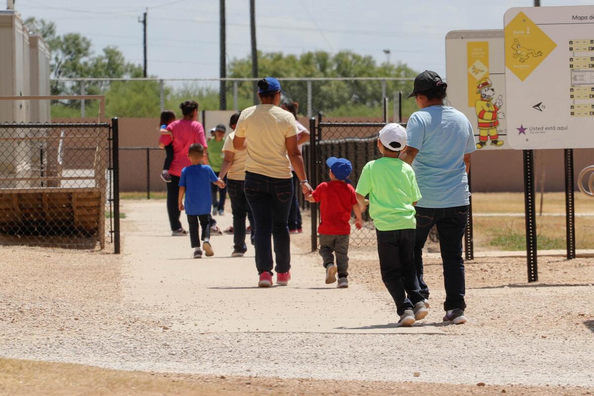 Adults hold hands with children as they walk from a dirt yard onto a sidewalk in a fenced area at an ICE detention center