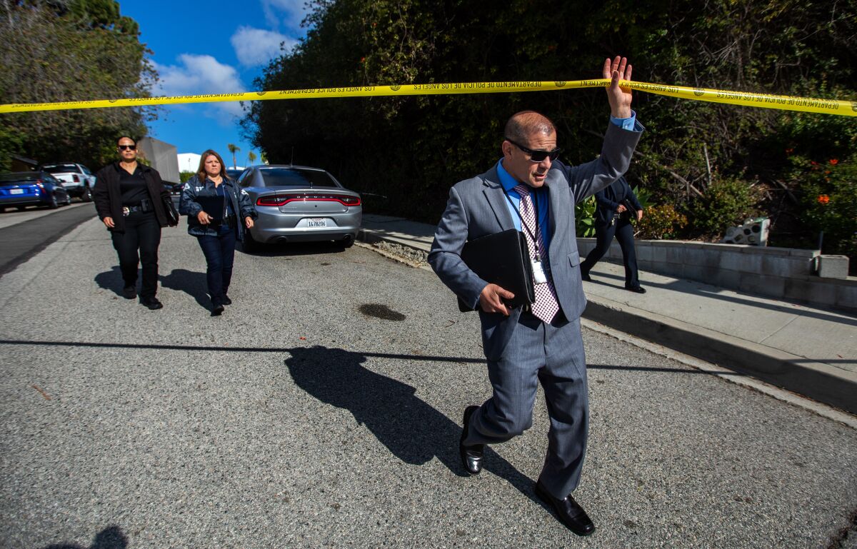 A man in a suit and sunglasses and a lanyard holds up crime scene tape as he walks beneath.