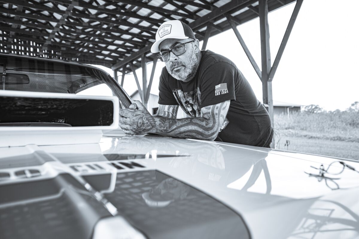 A man with tattoos and a trucker cap leans on the hood of a muscle car