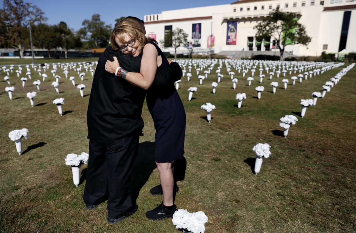 Two people embrace amid rows of white vases holding white flowers.