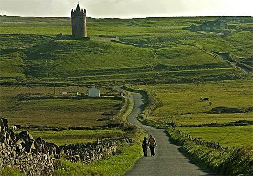A winding road leading to the sheer cliffs of Moher cuts through Ireland's emerald green countryside and is bordered by remnants of an ancient stone castle and fences.