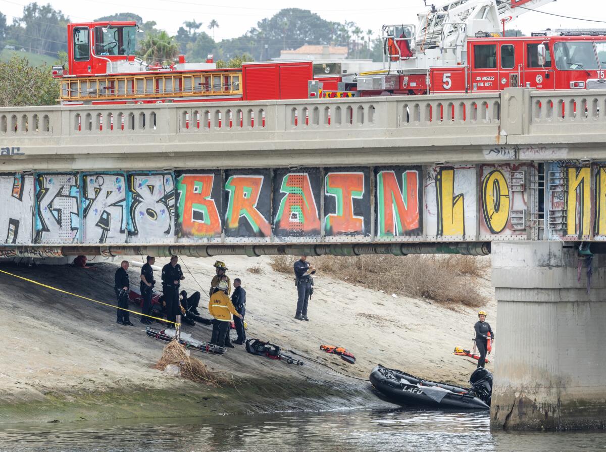 Firefighters in a concrete channel beneath a graffiti-tagged bridge, with firetrucks parked on the bridge.