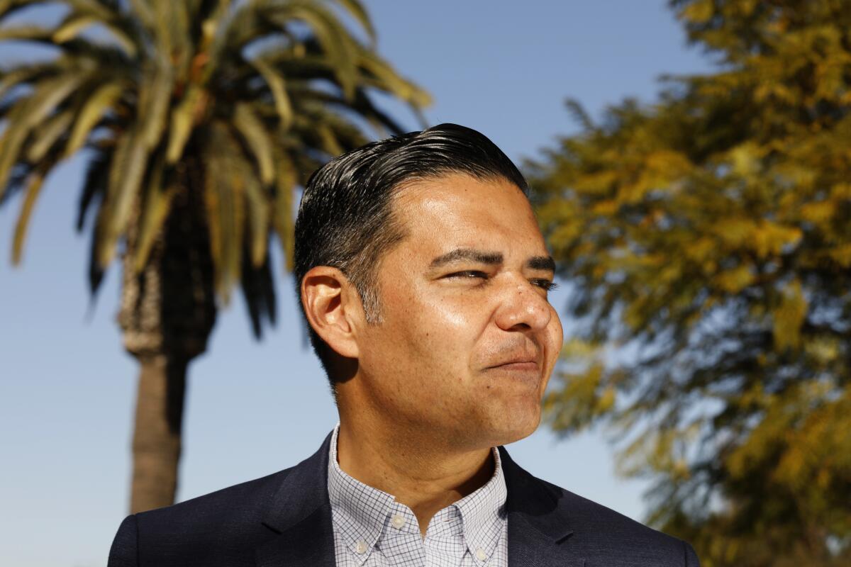 A man wearing a suit and a checked shirt stands in front of trees.