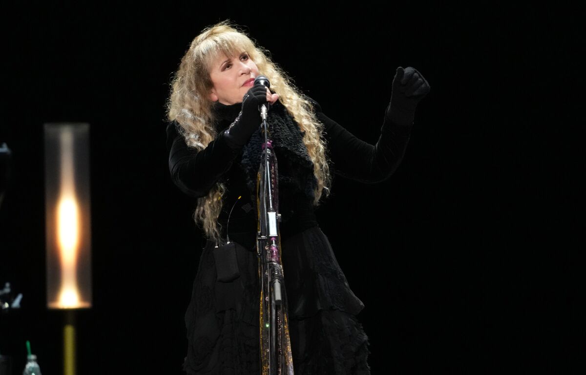 Stevie Nicks stands holding a microphone while dressed in black.