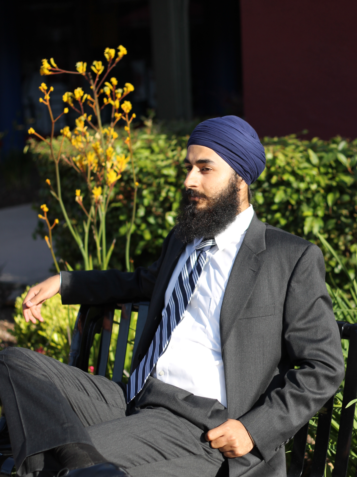 A man wearing a suit and tie and a blue turban sits in a chair outside.