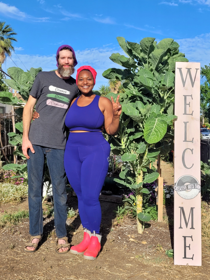 A man and a woman have their arms around each other, standing next to a tall plant and a vertical sign that says "welcome."