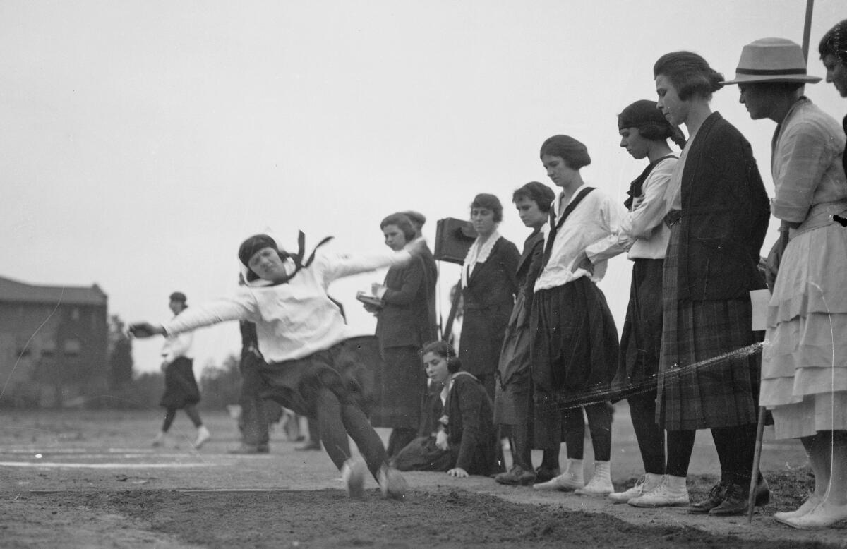 A young woman in a sailor top and bloomers lands in dirt after a long jump. 