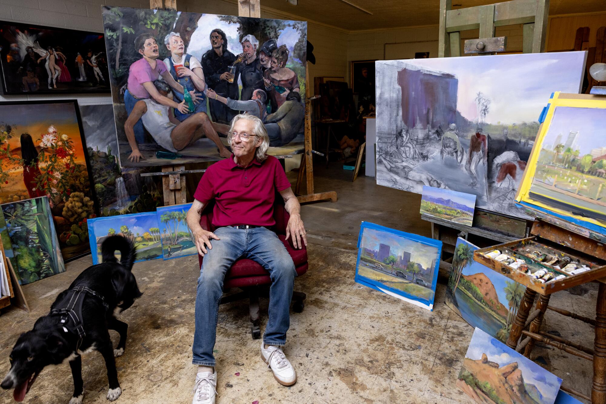 A man sits next to a dog surrounded by paintings in a studio