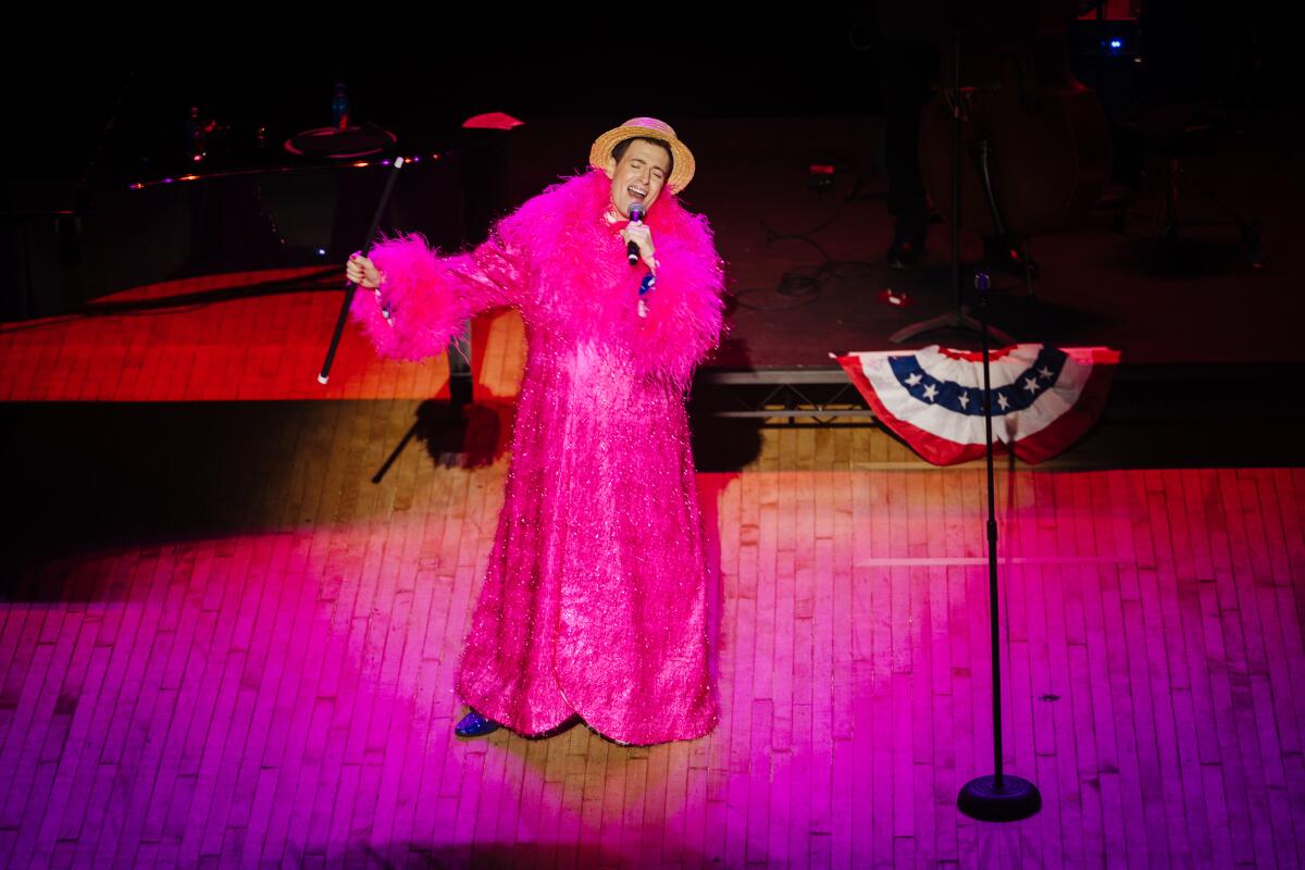 A man in a pink gown and a straw hat sings on stage.