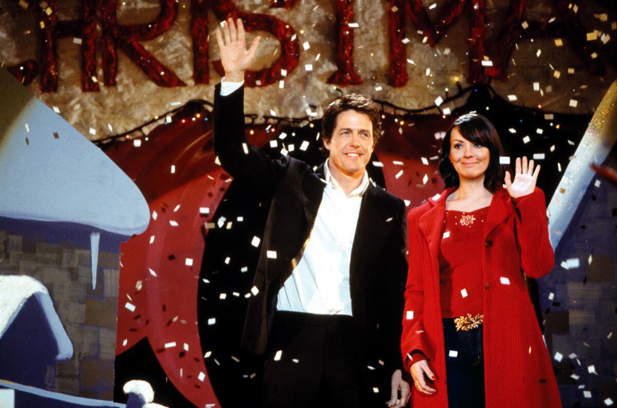 A man in a black suit and a woman in a red coat smile and wave as fake snow falls around them