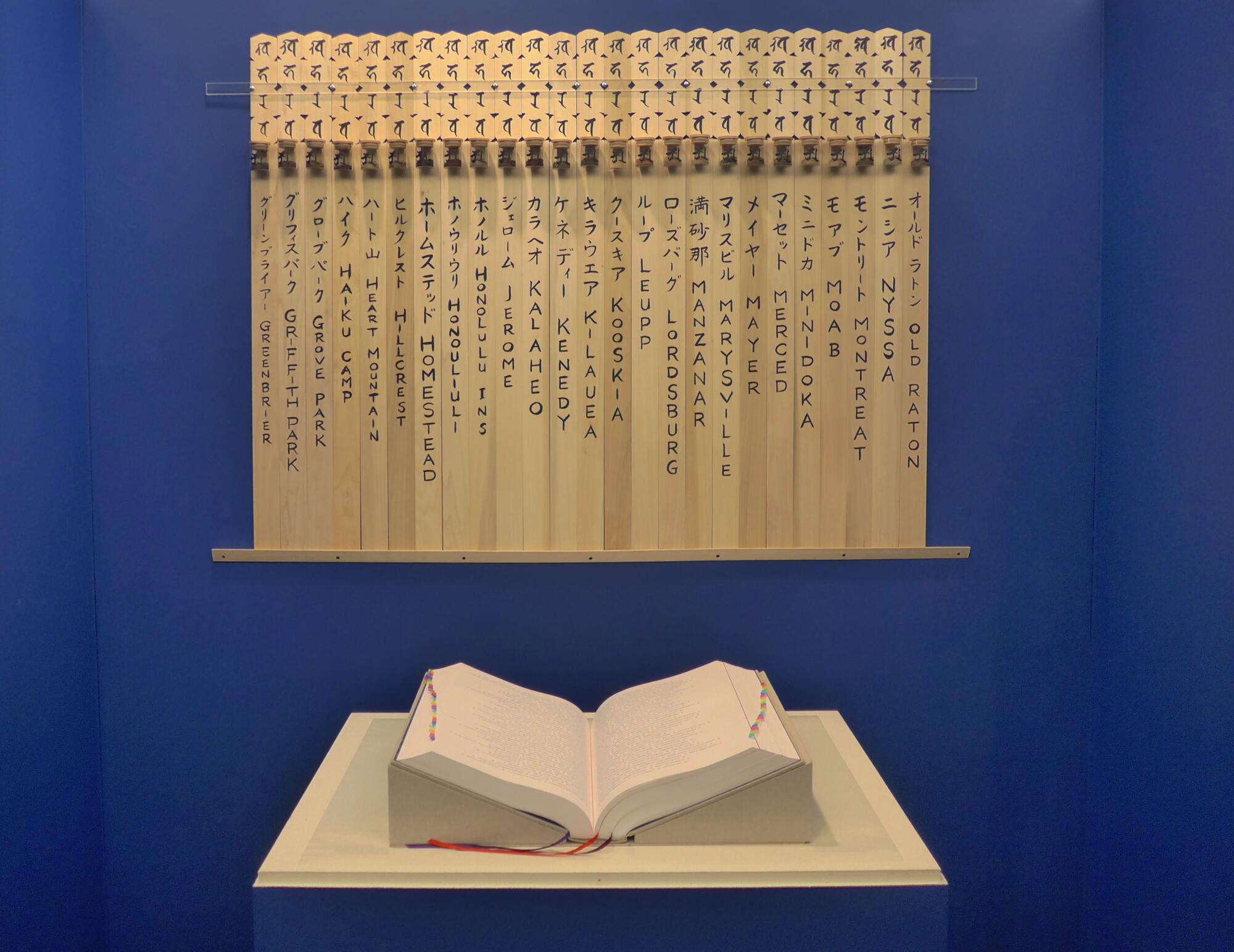 A large book is displayed open on a platform, below vertical wood planks with Japanese names.