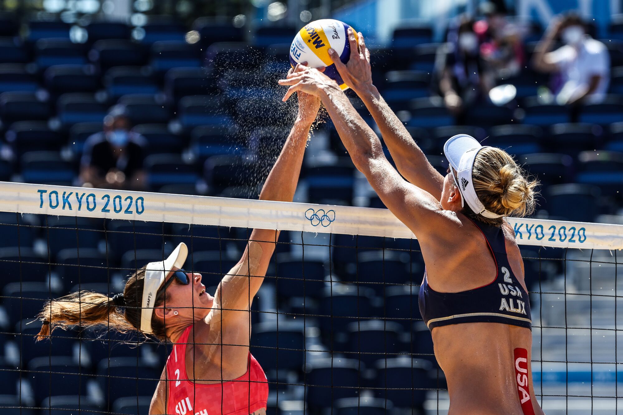 Two women leap up at the net with the volleyball between them.