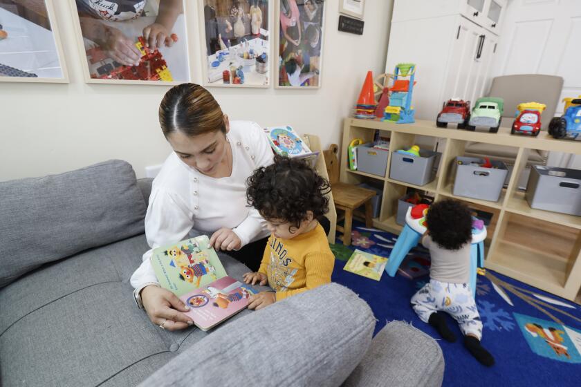 Lakewood, CA - May 17: Jennifer Cortez, childcare assistant, reads to Enzo Muniz, 20-months-old, while Luca Brown, 8 months old, plays at right, at Zoila Carolina Toma's family childcare center in Lakewood Wednesday, May 17, 2023. Zoila is licensed to care for up to 14 children from 8 months-12 years old Inside her center. They have a nap room, an art area, and a reading area to promote a comfortable atmosphere where students can engage in their activities. Currently, Zoila is at capacity, but she is constantly receiving calls from families looking for high-quality care. The need for care is desperately there, but there are not enough family child care centers to cover the needs of families, and few want to enter an industry where wages are so low. (Allen J. Schaben / Los Angeles Times)