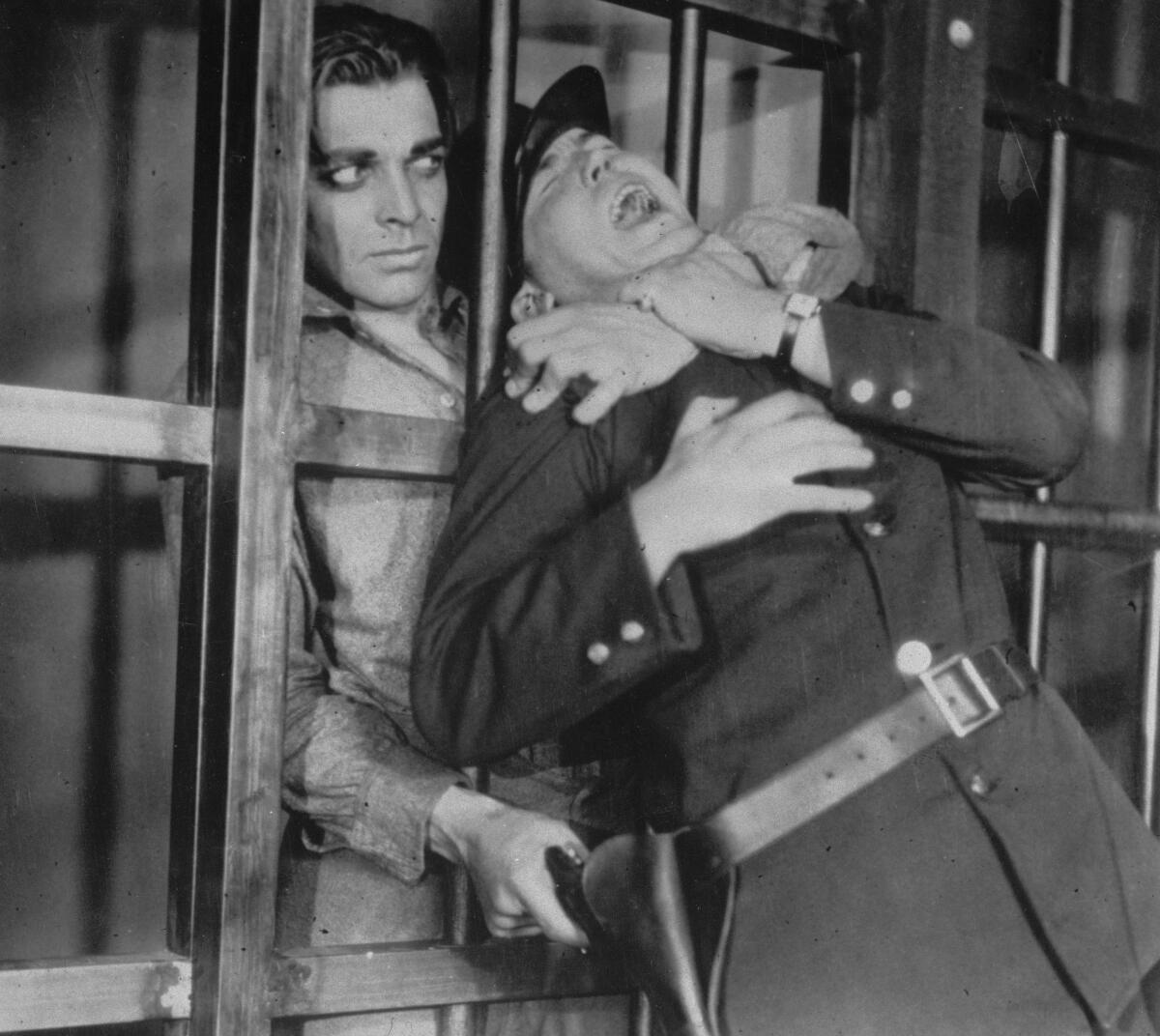 A man with dramatic eye makeup behind bars grabs the neck of another man in prison guard uniform and grabs for his gun.