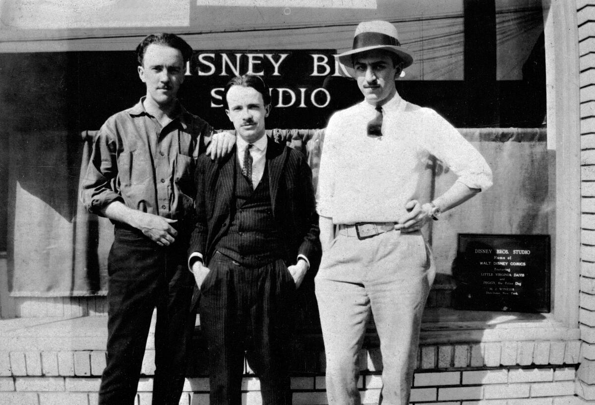 Three men stand in front of a sign that says Disney Brothers Studio.
